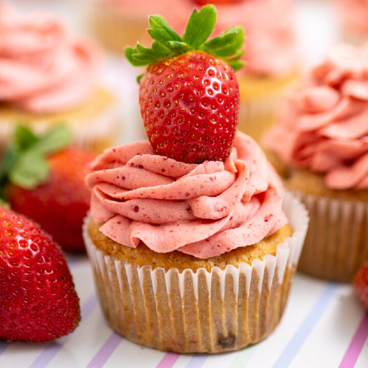Strawberry cupcake topped with strawberry frosting and a fresh strawberry, surrounded by more fresh strawberries and cupcakes on a pastel striped background.