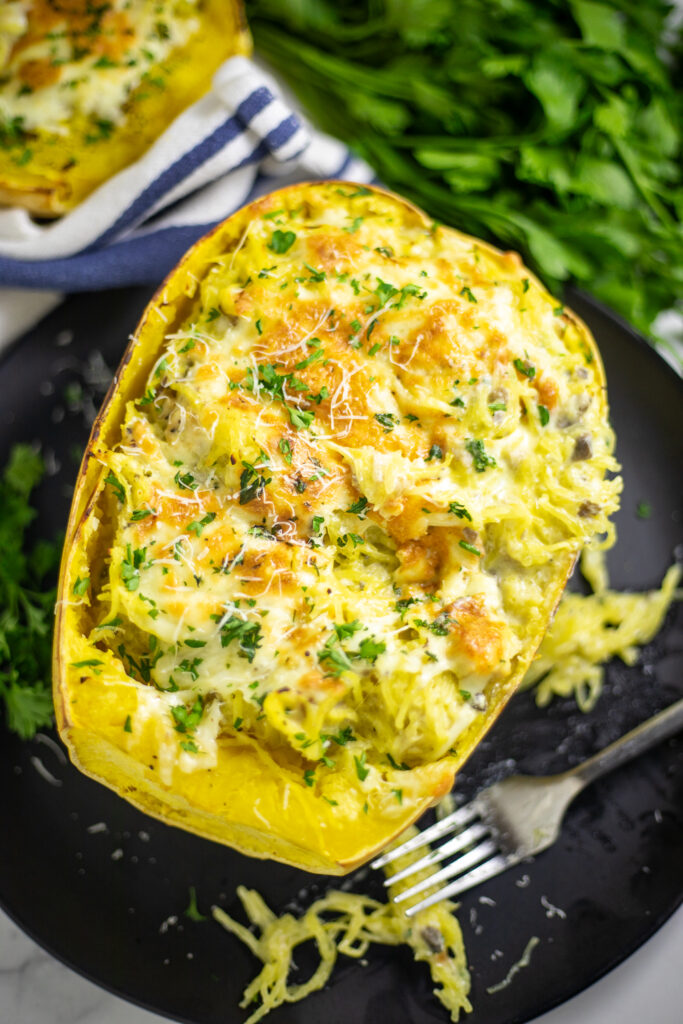 Overhead view of Alfredo spaghetti squash top with melted cheese and fresh parsley on a black plate next to a fork with parsley and a blue striped linen napkin in the background.