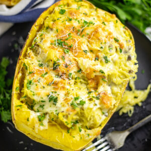 Overhead view of Alfredo spaghetti squash top with melted cheese and fresh parsley on a black plate next to a fork with parsley and a blue striped linen napkin in the background.