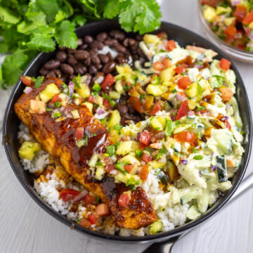 BBQ Salmon bowls with white rice, black beans, slaw, pineapple salsa, and barbecue salmon in a wide black bowl on a white wood background with fresh cilantro behind.