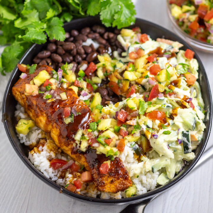 BBQ Salmon bowls with white rice, black beans, slaw, pineapple salsa, and barbecue salmon in a wide black bowl on a white wood background with fresh cilantro behind.