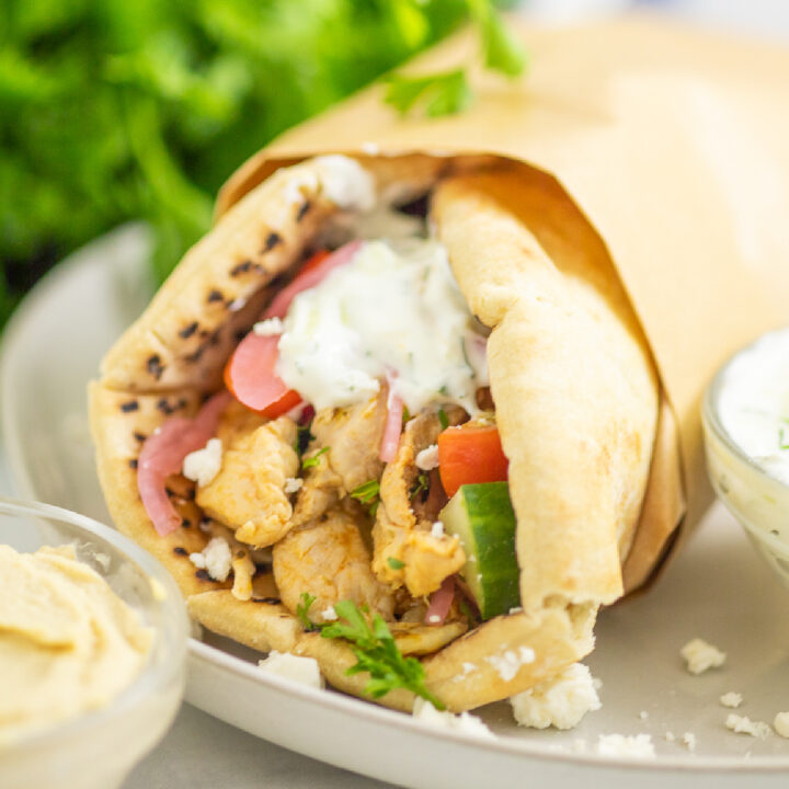 A chicken gyro topped with tzatziki on a white plate next to hummus with a bunch of fresh parsley behind.