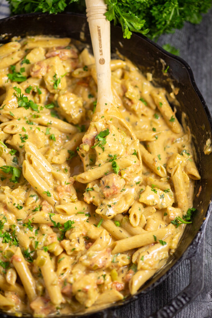 Overhead view of Creamy Cajun Chicken Pasta in a cast iron skillet topped with fresh parsley with a spoon in the middle. Skillet is on a dark rustic wood background next to a bunch of parsley.