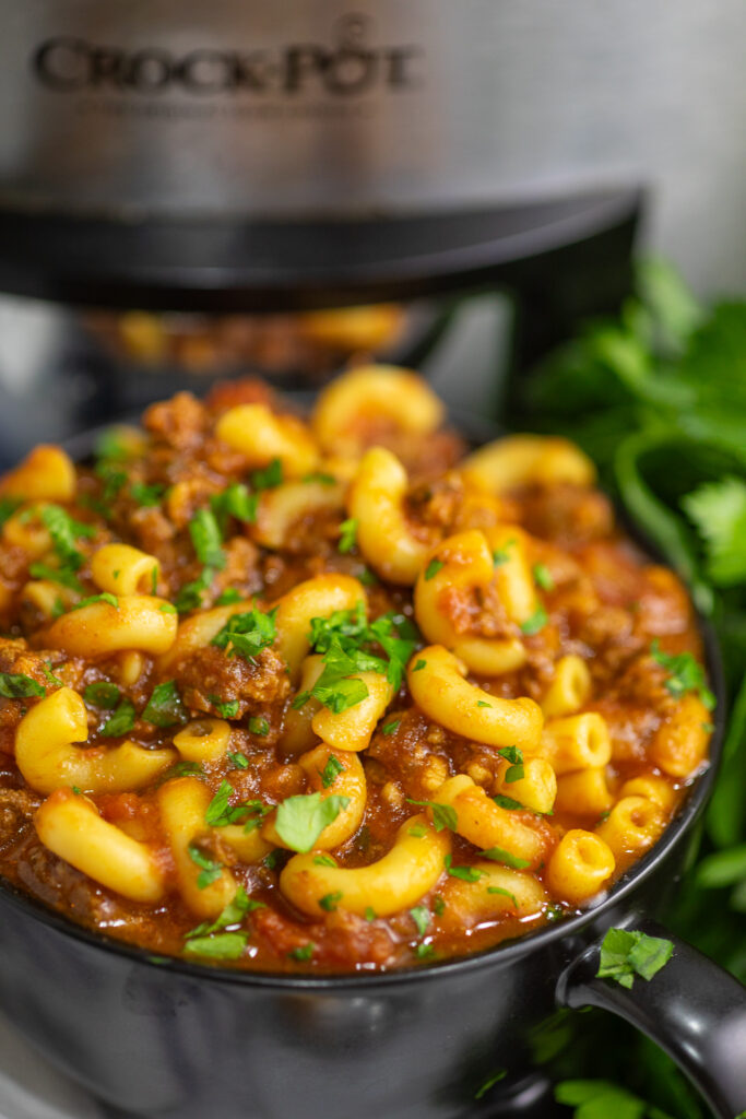 Crockpot goulash topped with fresh parsley in a black bowl with a bunch of fresh parsley and a stainless steel CrockPot behind.