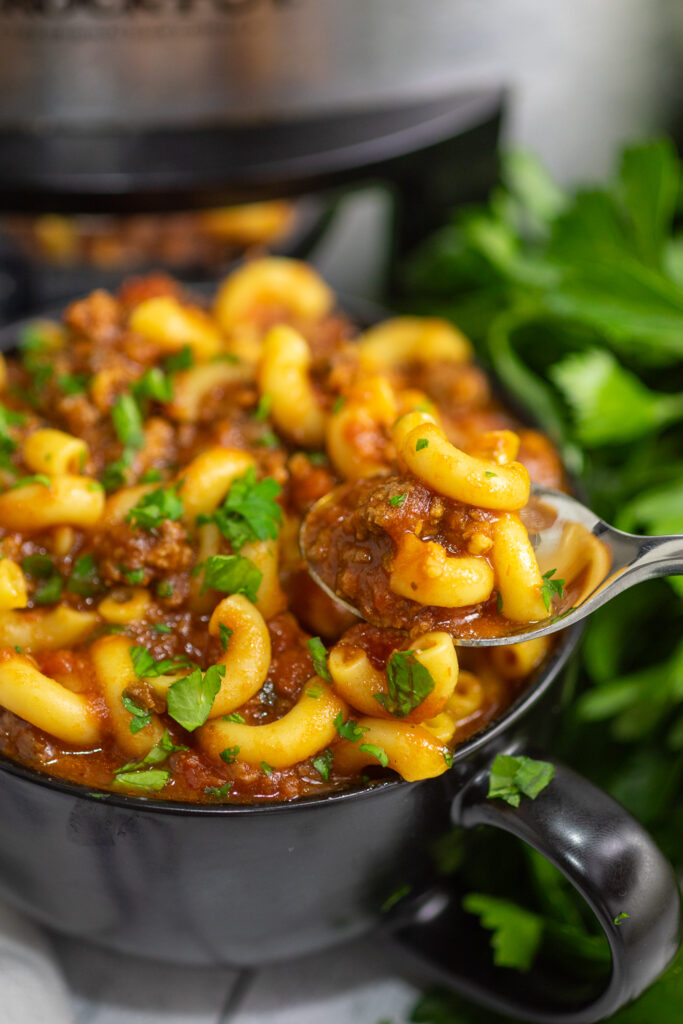 A spoon taking a scoop of crockpot goulash from a black bowl with a bunch of fresh parsley and a stainless steel CrockPot behind.