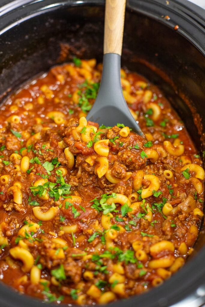 Overhead close up view of crockpot goulash in a slow cooker topped with fresh parsley with a wooden spoon in the middle.