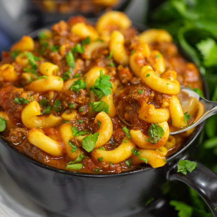 A black bowl full of crockpot goulash topped with fresh parsley with a metal spoon taking a bite out and parsley behind.