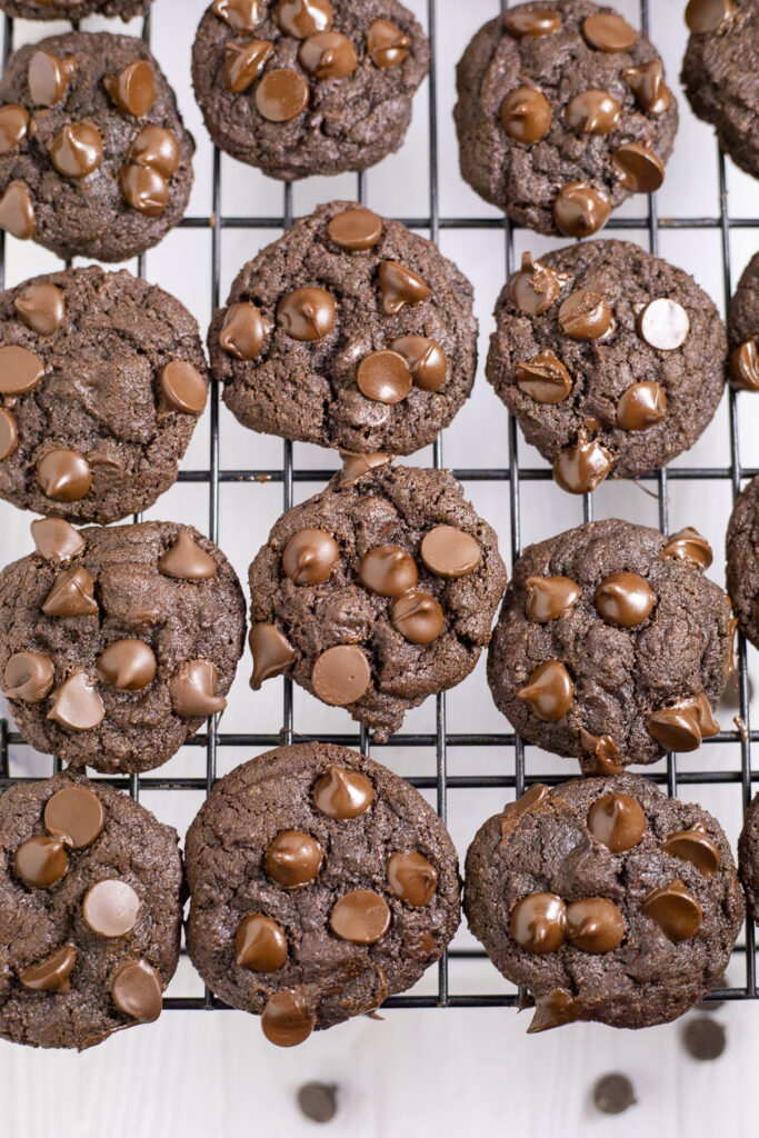 Overhead view of double chocolate chip cookies on a cooling rack over a white background with chocolate chips scattered around.