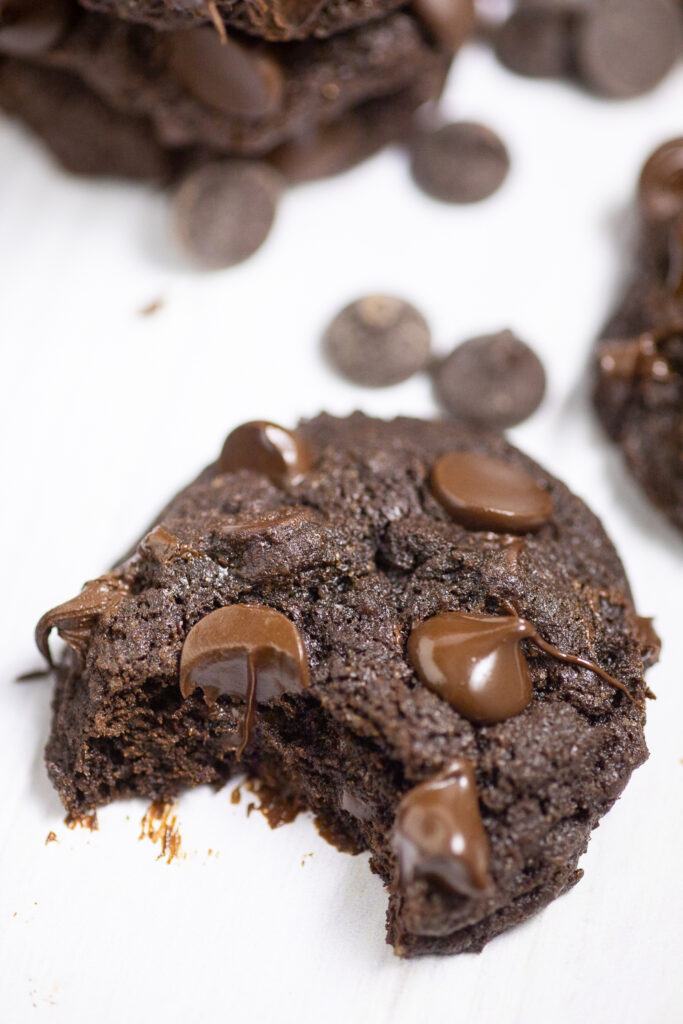 A double chocolate chip cookie with a bite taken out on a white background with more cookies and large chocolate chips behind.