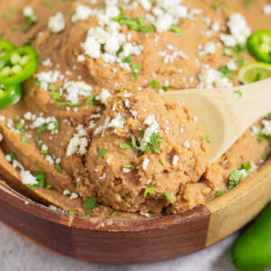 Wooden spoon scooping up a big serving of instant pot refried beans from a wooden bowl topped with cilantro and crumbled cotija.