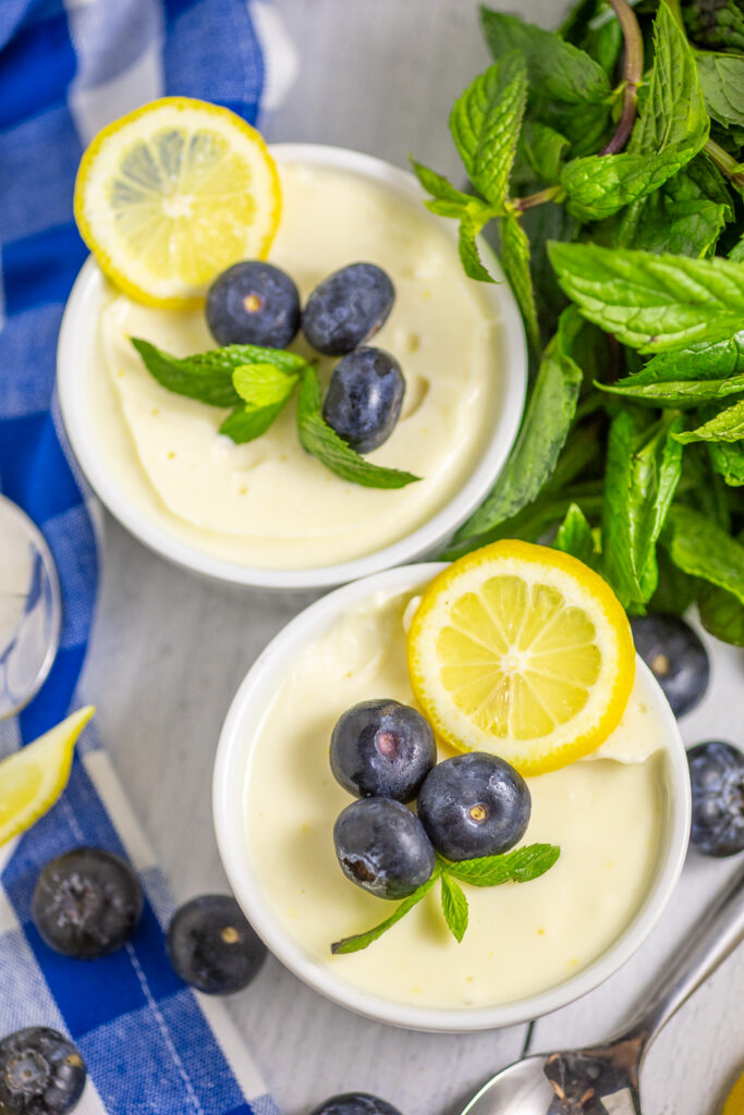 Overhead view of two white ramekins filled with lemon posset and topped with fresh blueberries, mint, and a lemon slice. The ramekins are next to more fresh mint, a bright blue plaid napkin, and fresh blueberries scattered around.
