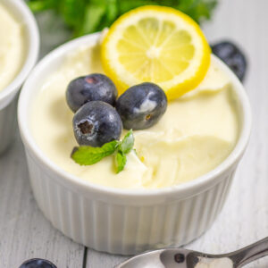 Lemon posset in a white ramekin with a bite missing topped with three fresh blueberries, a small sprig of mint, and a lemon slice. The ramekin is on a white rustic wood background with a second ramekin and more mint behind.