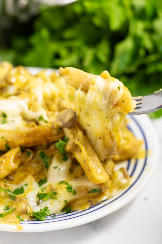 A fork picking up a cheesy bite of Philly Cheesesteak Pasta from a small white plate with a bunch of parsley behind.