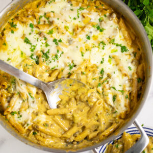 Overhead view of a pot of Philly Cheesesteak pasta topped with melted cheese and fresh parsley with a scoop taken out and a metal spoon in the middle.