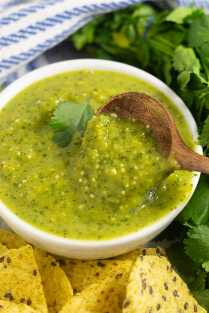A small white bowl filled with homemade salsa verde with a wooden spoon taking a scoop out, with chips surrounding the bowl and a bunch of fresh cilantro and blue and white striped linen napkin behind.