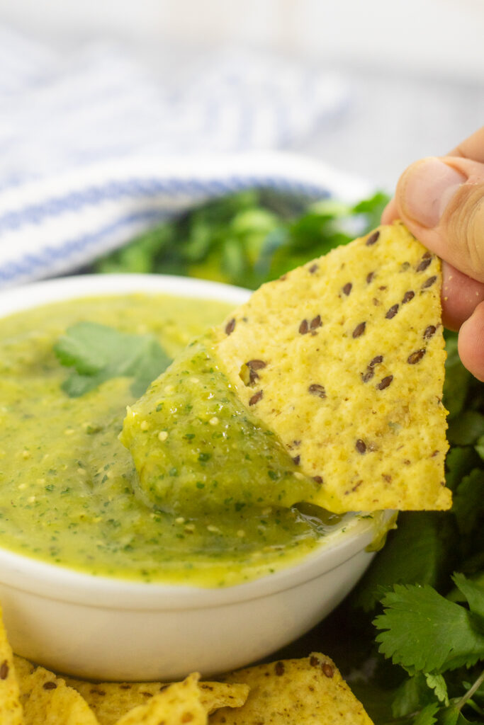 A tortilla chip dipping into a small white bowl of salsa verde with chips surrounding the bowl and a bunch of fresh cilantro and blue and white striped linen napkin behind.