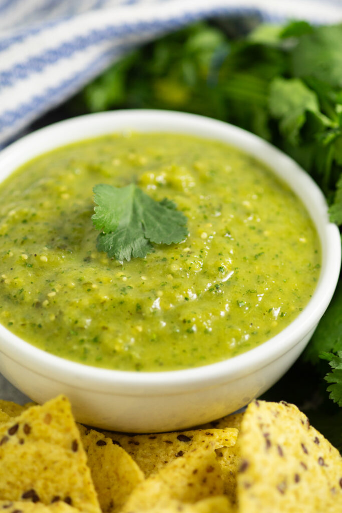 A small white bowl filled with salsa verde and topped with a leaf of fresh cilantro, surrounded by tortilla chips, fresh cilantro, and a blue and white striped linen napkin.