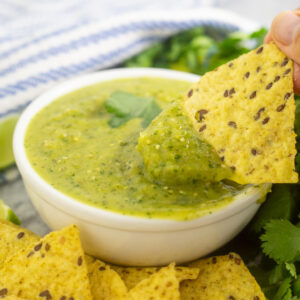 A tortilla chip dipping into a small white bowl of salsa verde with chips surrounding the bowl and a bunch of fresh cilantro and blue and white striped linen napkin behind.