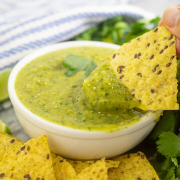 A tortilla chip dipping into a small white bowl of salsa verde with chips surrounding the bowl and a bunch of fresh cilantro and blue and white striped linen napkin behind.