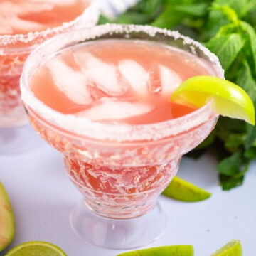 Watermelon margarita on the rocks in a chilled glass rimmed with salt and garnished with a lime wedge. There's a second margarita and fresh mint behind.