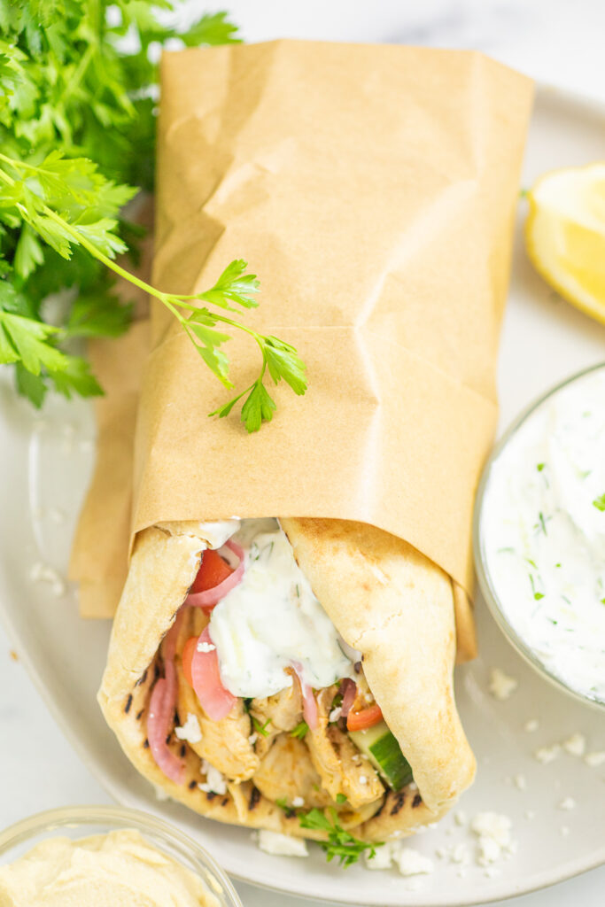 Overhead view of a chicken gyro wrapped in brown paper on a white plate, topped with pickled onions and tzatziki. The gyro sits next to a small bowl of tzatziki, a lemon wedge, and a bunch of fresh parsley.