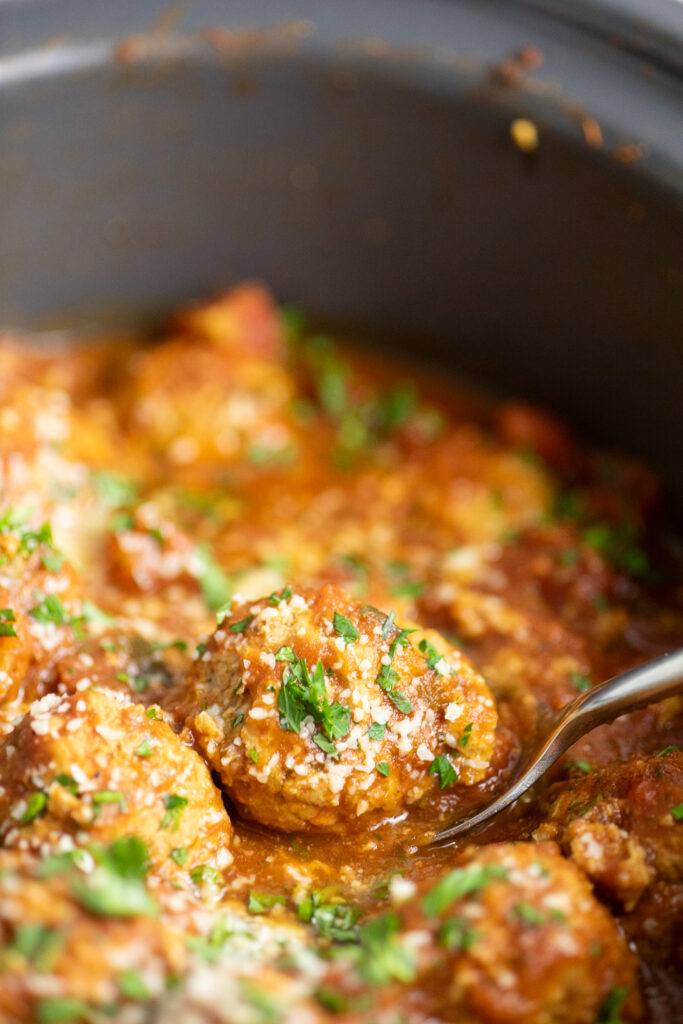 A metal spoon scooping a turkey meatball in red sauce from a slow cooker.