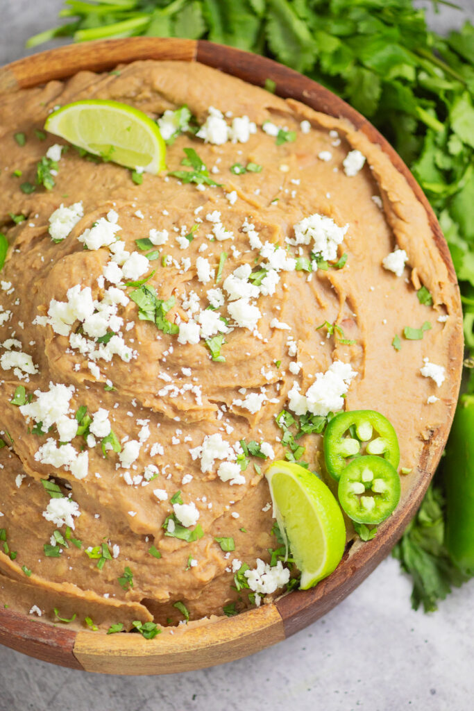 Overhead view of Instant Pot Refried beans topped with cilantro, crumbled cotija, and lime wedges with a bunch of fresh cilantro behind.