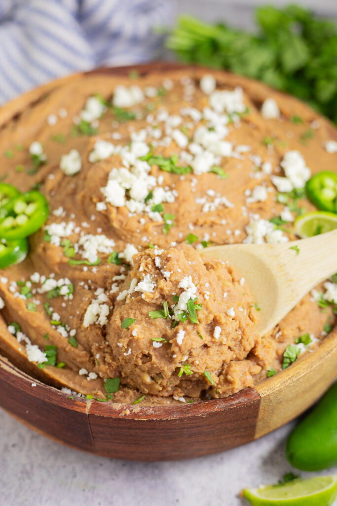 Wooden spoon scooping up a big serving of instant pot refried beans from a wooden bowl topped with cilantro and crumbled cotija.