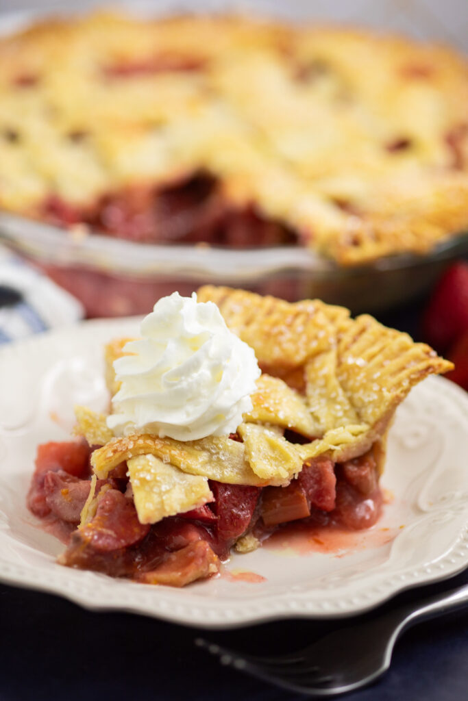 A slice if strawberry rhubarb pie on a small white plate topped with a swirl of whipped cream with the rest of the pie behind in a glass dish.