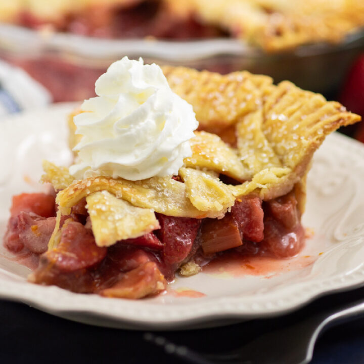 A slice if strawberry rhubarb pie on a small white plate topped with a swirl of whipped cream with the rest of the pie behind in a glass dish.