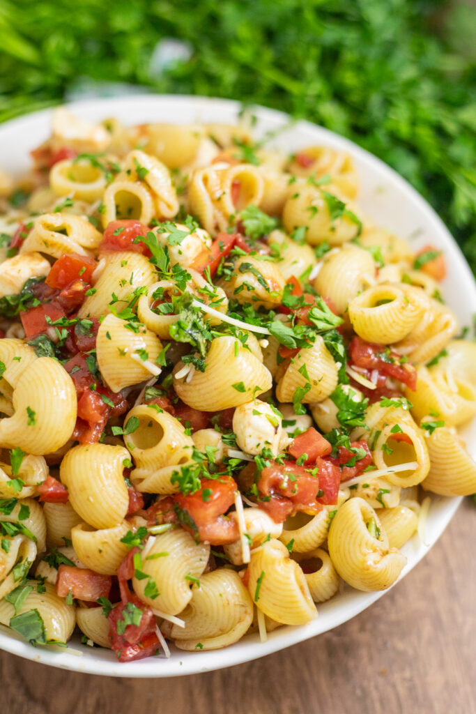 Overhead view of Bruschetta pasta salad in a wide white bowl on a wood table with a cloth linen and a bunch of fresh parsley behind.