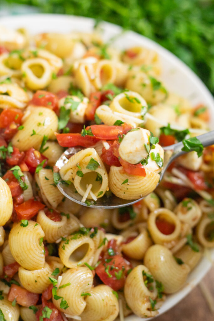 A close up view of a metal spoon taking a scoop of bruschetta pasta salad topped with freshly chopped parsley in a bowl.
