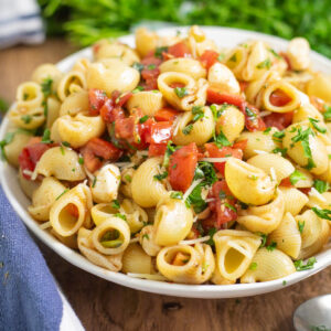 Bruschetta pasta salad in a wide white bowl on a wood table with a cloth linen and a bunch of fresh parsley behind.
