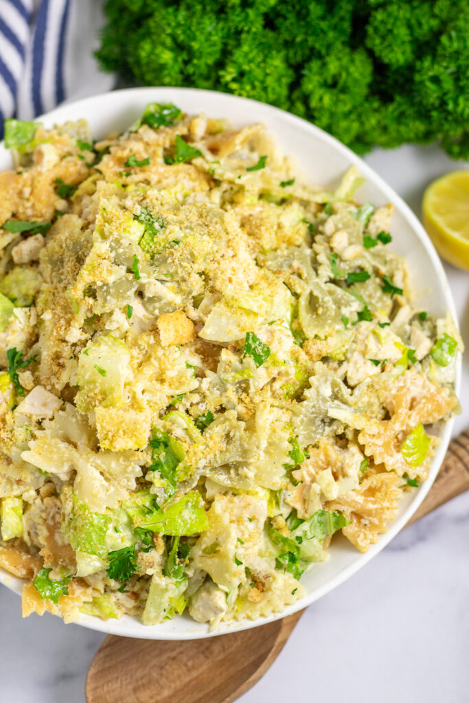 Overhead view of chicken caesar pasta salad in a white bowl topped with shredded parmesan cheese. The bowl is next to a wooden spoon and a bunch of fresh parsely on a white background.
