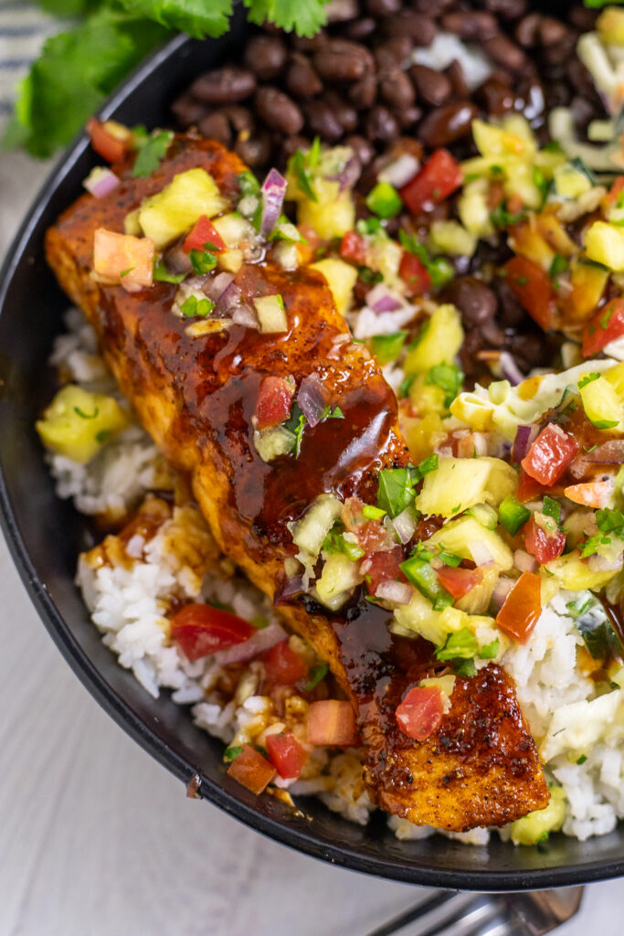 Close up of barbecue salmon in a BBQ salmon bowl topped with pineapple salsa with black beans and white rice in a black bowl on a white wood background.