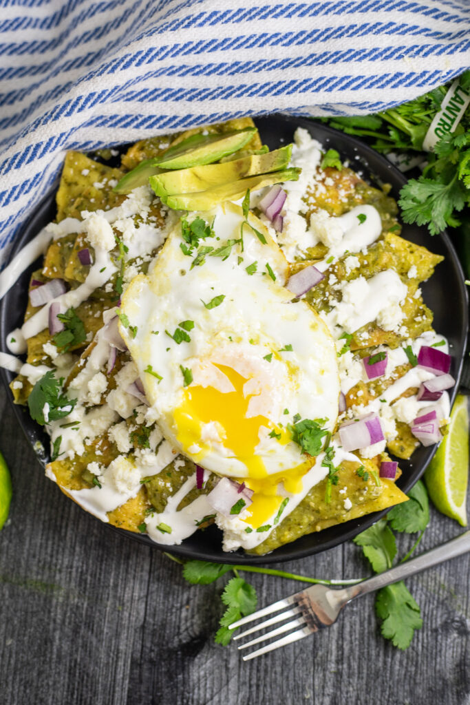 Overhead view of chilaquiles verdes topped with crema, red onion, avocado, cilantro, and a fried egg with the yolk running down on a black plate. The plate is on a rustic wood background with a blue striped linen napkin and a fork.