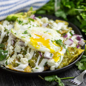 Chilaquiles verdes topped with crema, red onions, cilantro, and a fried egg with the yolk running down. There is a striped linen napkin and a fresh bunch of cilantro behind.