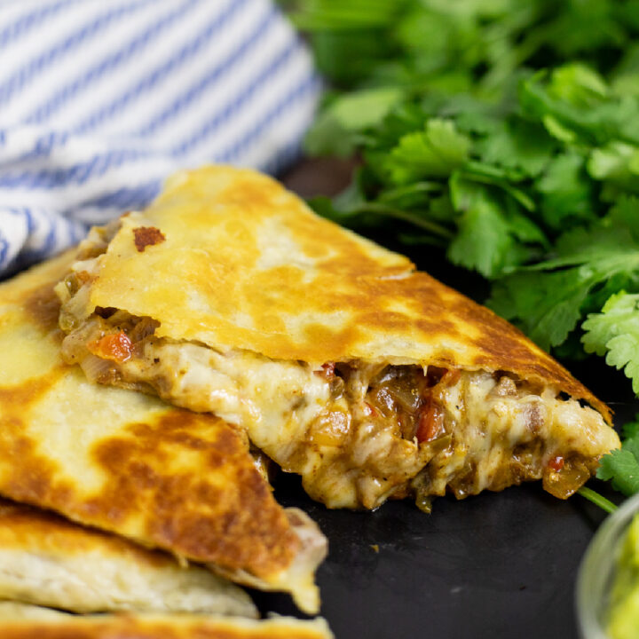 Side view of a fajita quesadilla wedge with gooey melted cheese oozing from the side and a striped linen napkin and a bunch of fresh cilantro behind.