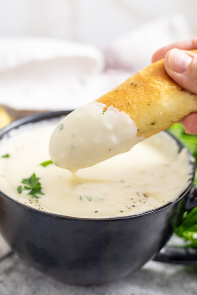 A garlic breadstick dipping into a small black bowl full of homemade alfredo sauce with more breadsticks behind.