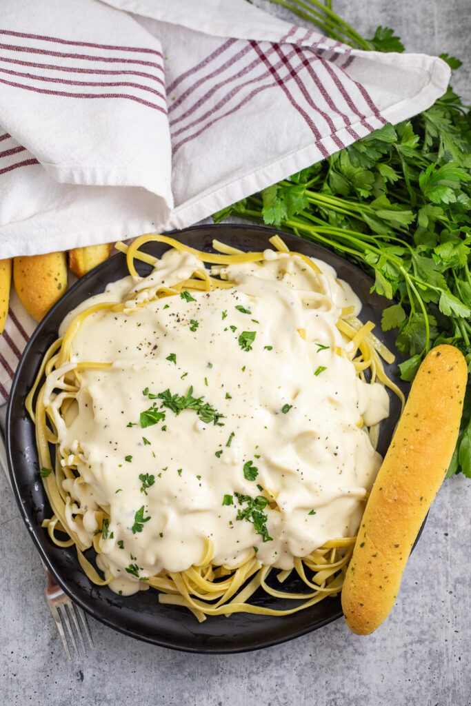 Overhead view of fettuccine noodles on a large black plate smothered in homemade Olive Garden Alfredo sauce, next to a garlic breadstick and topped with fresh chopped parsley. There are more breadsticks wrapped in a red and white linen napkin and a fresh bunch of parsley behind the plate on a gray stone background.