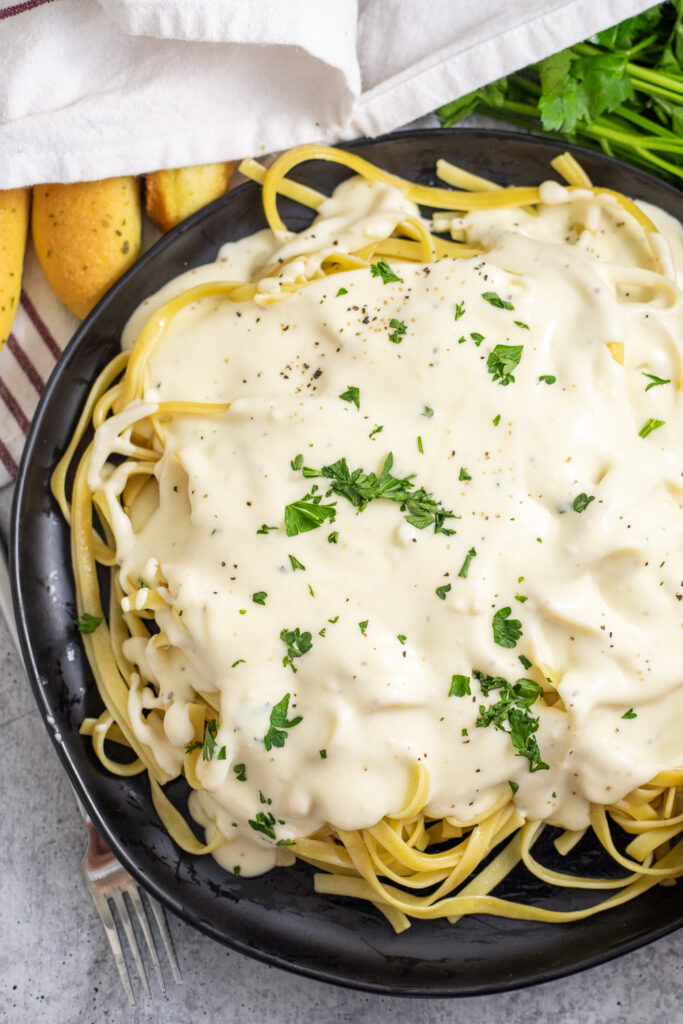 Close up of homemade Olive Garden Alfredo Sauce on top of fettuccine noodles next to a breadstick.