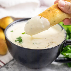 A garlic breadstick dipping into a small black bowl full of homemade alfredo sauce with more breadsticks behind.