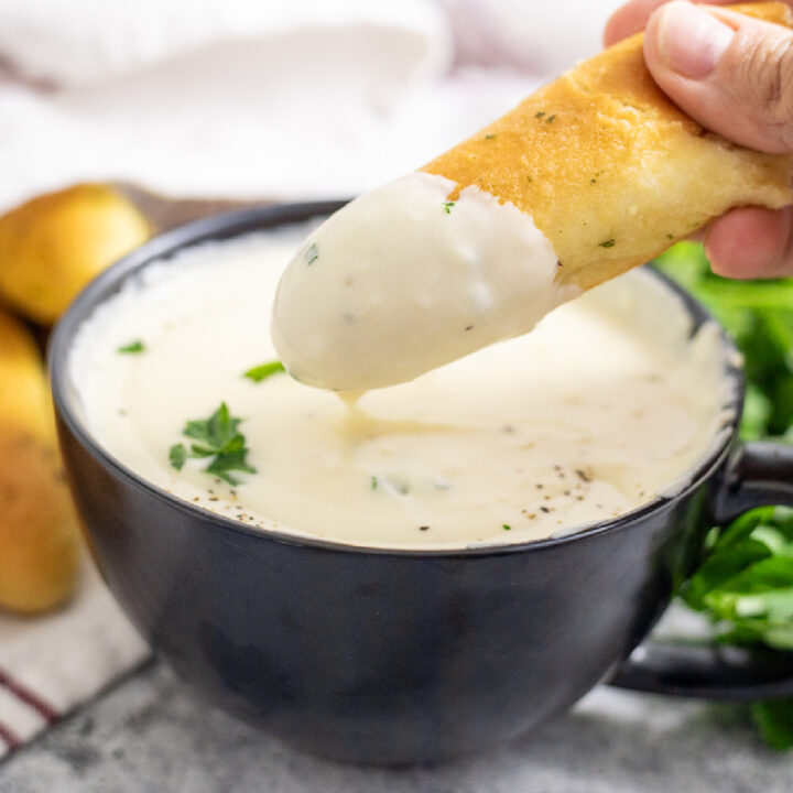 A garlic breadstick dipping into a small black bowl full of homemade alfredo sauce with more breadsticks behind.