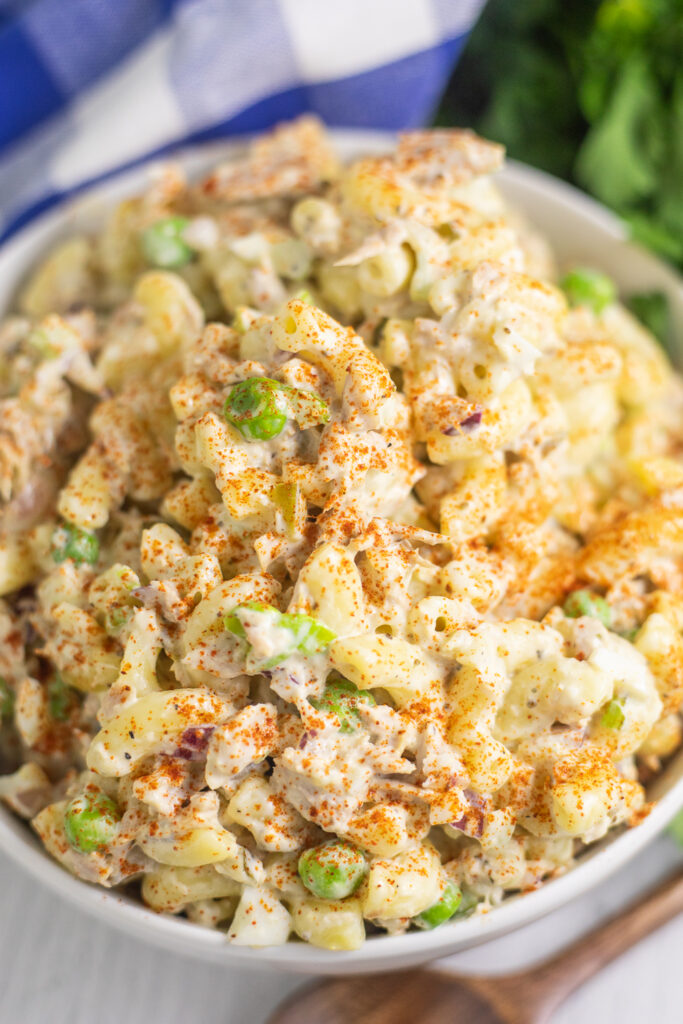 Overhead view of a white bowl of tuna macaroni salad topped with smoked paprika with a blue checkered linen behind.