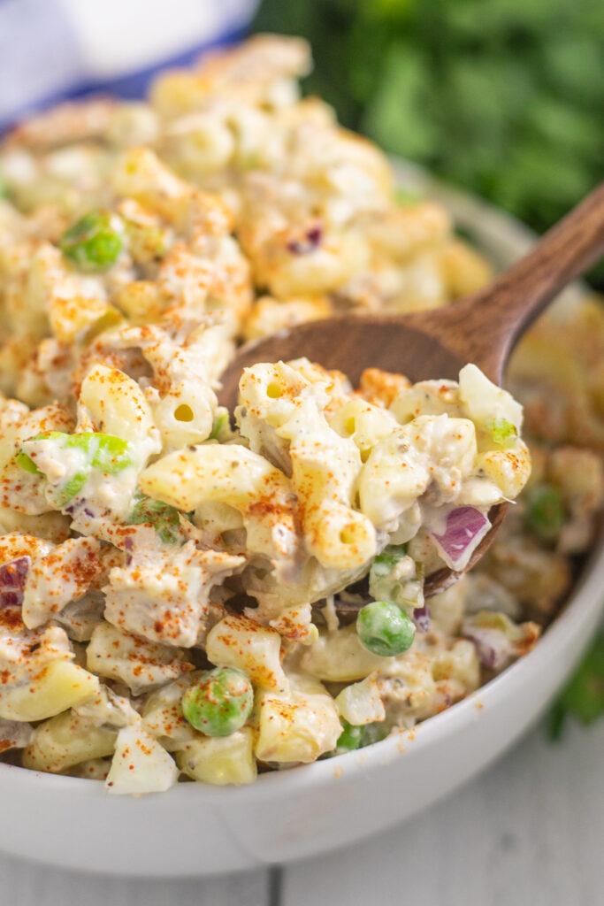 Tuna macaroni salad in a wide white bowl with a wooden spoon sticking out and a blue checkered linen napkin and fresh bunch of parsley behind on a white wooden plank background.