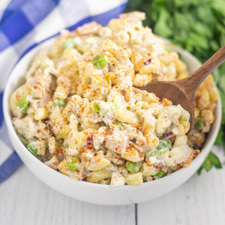 Tuna macaroni salad in a wide white bowl with a wooden spoon sticking out and a blue checkered linen napkin and fresh bunch of parsley behind on a white wooden plank background.