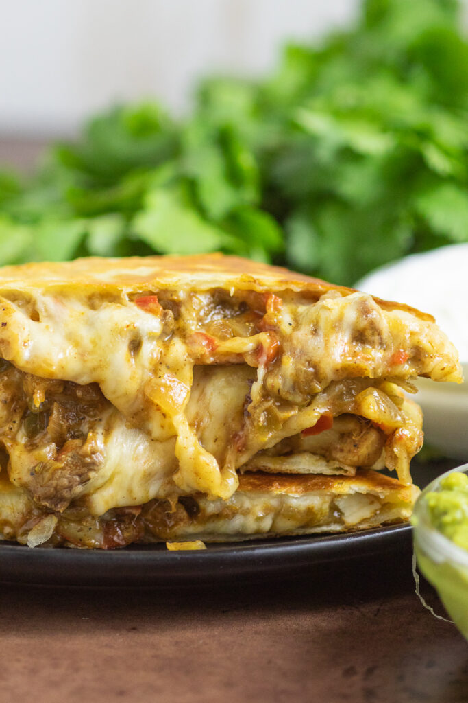 Fajita Quesadilla triangles stacked on top of each other with melty cheesy filling with beef, chicken, onion, and peppers showing and dripping down. The quesadilla is on a black plate with a bunch of fresh cilantro behind.