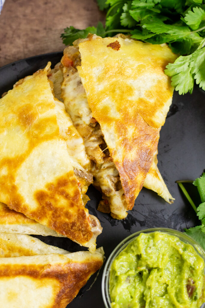 Overhead view of a fajita quesadilla with a sneak peak of the cheesy beef, chicken, and pepper filling, next to a small glass bowl of guacamole and a sprig of fresh cilantro.