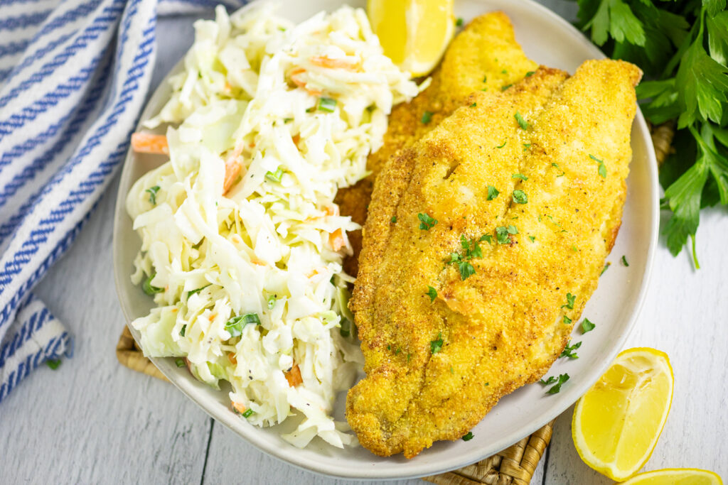 Air fryer catfish with cornmeal breading on a beige ceramic plate next to a large serving of coleslaw, surrounded by a striped linen napkin and fresh parsley on a white wood background.