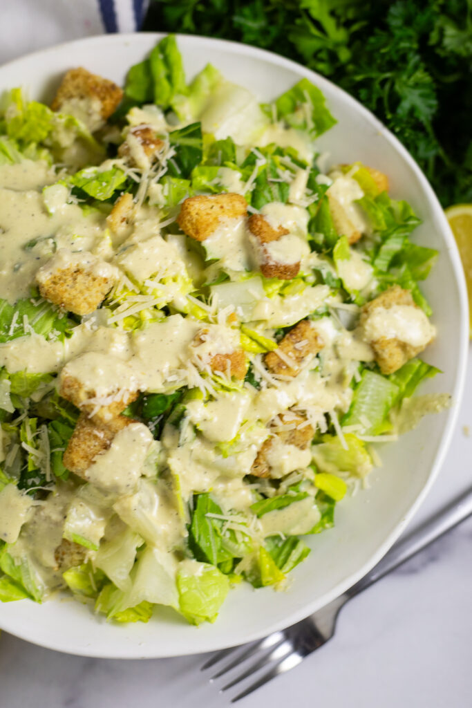 Overhead view of a Caesar salad topped with homemade caesar dressing, shredded parmesan, and croutons in a wide white bowl on a white background next to a fork and fresh parsley.